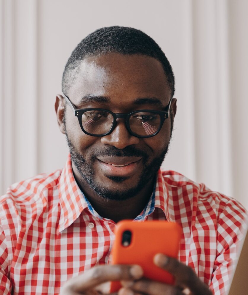 Smiling African ethnicity man office worker sitting at desk holding smartphone chatting with friends